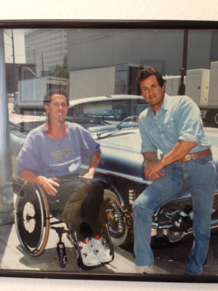 Two men sitting in front of a car and one man is in a wheelchair.